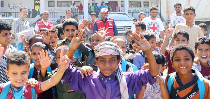 Photographie d'enfants entre 6 et 12 ans souriants et levant les bras en premier plan. Derrière eux, des personnes travaillant pour la Croix-Rouge regardent la scène.
