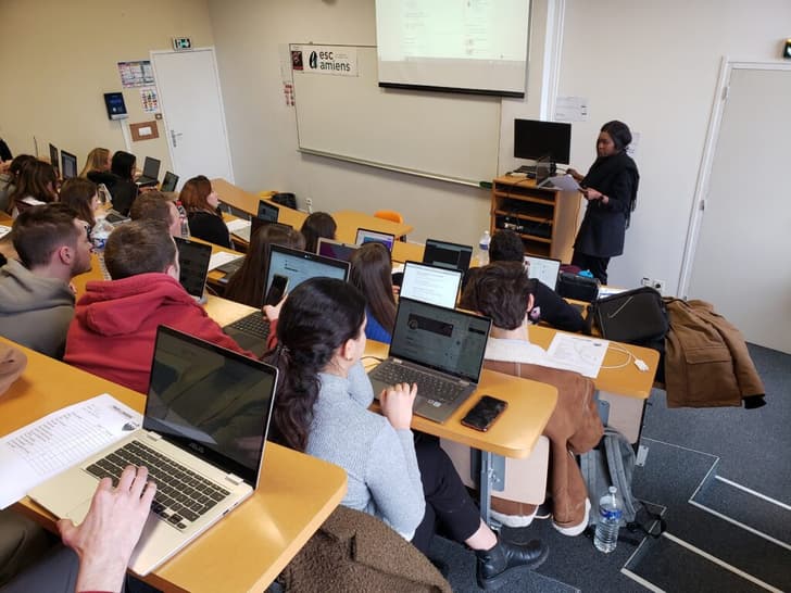 Photographie d'un cours donné. On aperçoit des étudiants prenant des notes sur leurs ordinateurs, et un professeur en arrière-plan.