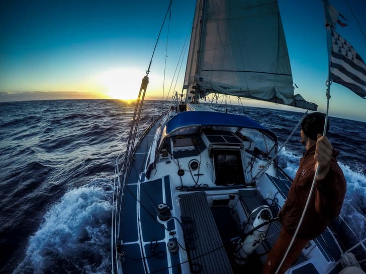 Photographie du bateau Maewan en pleine mer. Un homme debout se tient à une corde où flotte un drapeau breton. L'homme regarde la mer agitée, et le soleil au loin en train de se coucher.