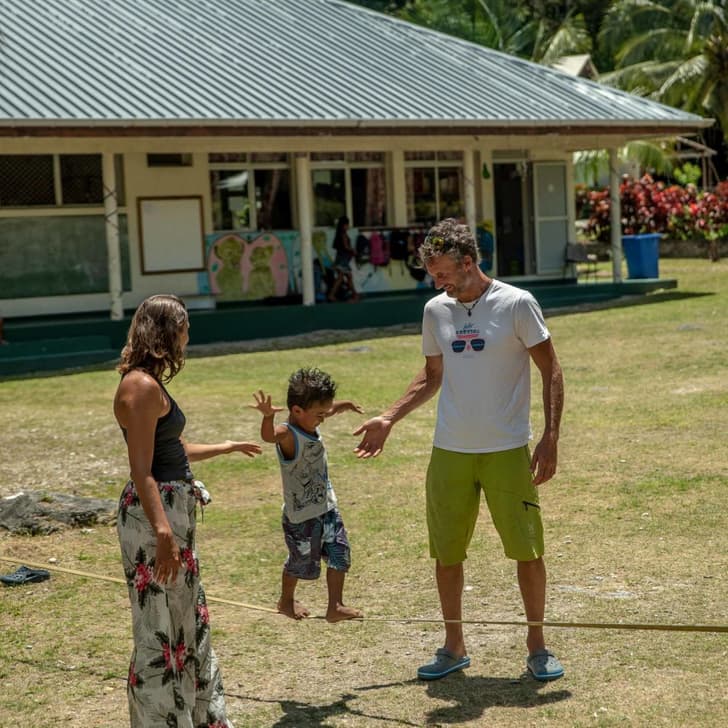 Photographie d'un jeune enfant en train de marcher en équilibre sur une slackline. À ses côtés se trouve Marion Courtois et Erwan Le Lann, les deux leaders de l'expédition, prêts à aider l'enfant. En fond, nous pouvons apercevoir une école avec des dessins d'enfants sur les murs.